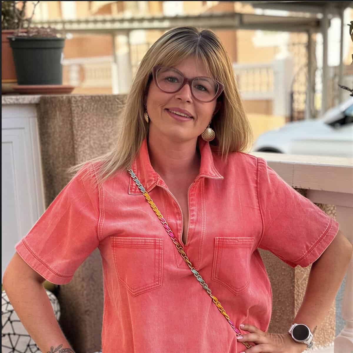 Vrouw met blonde haar en roze blouse poseert buiten op terras.